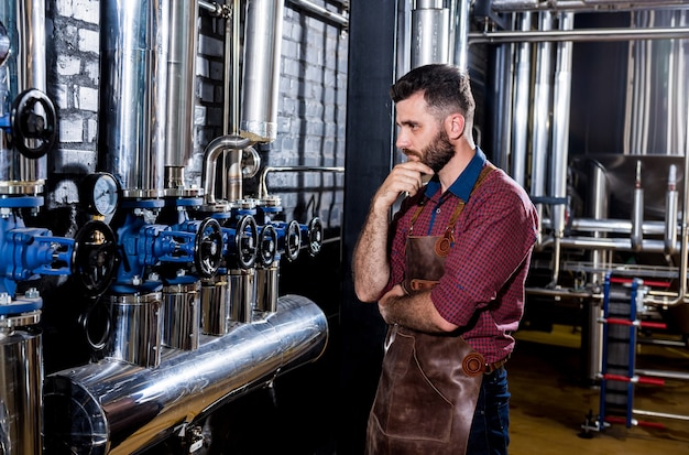 Giovane birraio maschio in grembiule in pelle che supervisiona il processo di fermentazione della birra nella moderna fabbrica di birra fa...