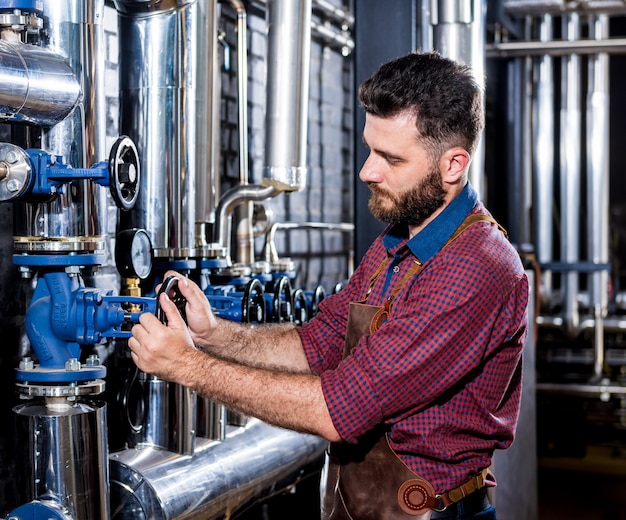 Giovane birraio maschio in grembiule in pelle che supervisiona il processo di fermentazione della birra nella moderna fabbrica di birra fa...