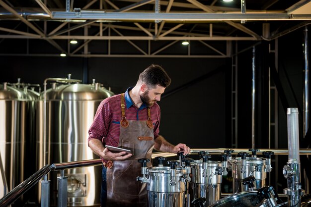 Giovane birraio maschio in grembiule in pelle che supervisiona il processo di fermentazione della birra presso il moderno birr...