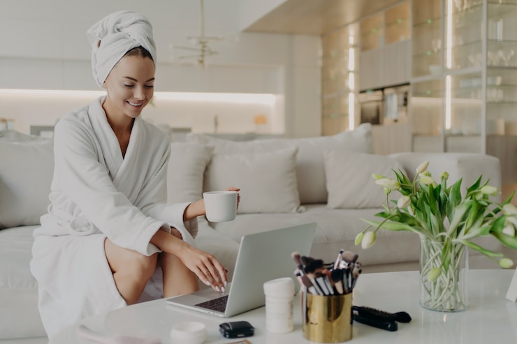 Foto giovane blogger di bellezza donna felice in accappatoio e asciugamano sulla testa seduto su un comodo divano nel soggiorno moderno e lavorando sul computer portatile, bevendo tè e sentendosi rilassato dopo aver fatto la doccia a casa