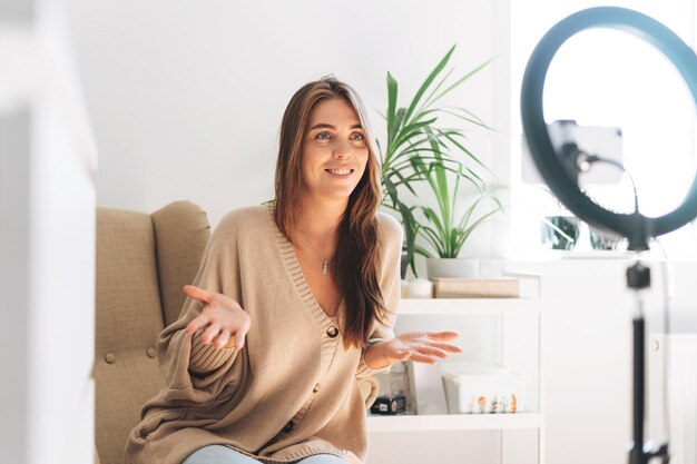 Giovane blogger donna graziosa in cardigan accogliente registra video sul telefono cellulare utilizzando una lampada ad anello seduta su una sedia a casa