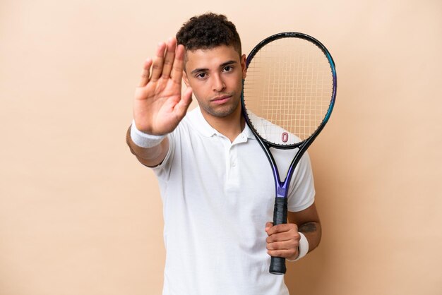 Giovane brasiliano bell'uomo che gioca a tennis isolato su sfondo beige che fa il gesto di arresto