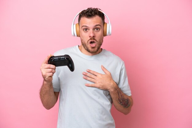 Giovane brasiliano che gioca con il controller per videogiochi isolato su sfondo rosa sorpreso e scioccato mentre guarda a destra