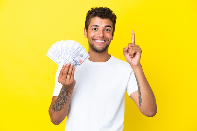 Giovane brasiliano che prende un sacco di soldi isolato su sfondo giallo che mostra e alza un dito in segno del meglio