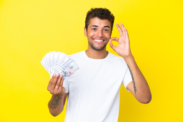 Giovane brasiliano che prende un sacco di soldi isolato su sfondo giallo che mostra il segno giusto con le dita