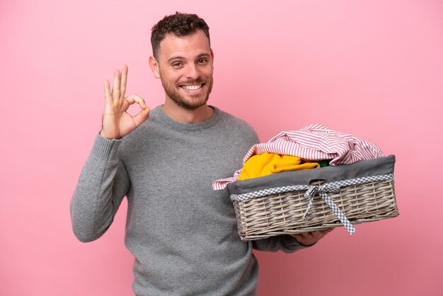 Giovane brasiliano che tiene un cesto di vestiti isolato su sfondo rosa che mostra il segno giusto con le dita