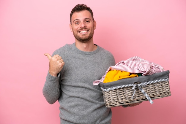 Giovane brasiliano che tiene un cesto di vestiti isolato su sfondo rosa che punta di lato per presentare un prodotto