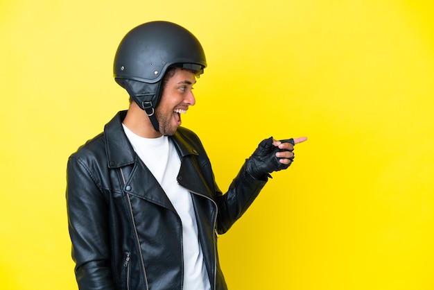 Giovane brasiliano con un casco da motociclista isolato su sfondo giallo che punta il dito di lato e presenta un prodotto