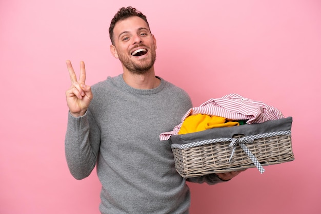 Giovane brasiliano in possesso di un cesto di vestiti isolato su sfondo rosa sorridente e mostrando il segno di vittoria