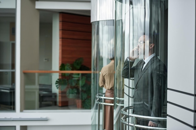 Giovane broker maschio in giacca e cravatta che parla con un collega sullo smartphone in ascensore