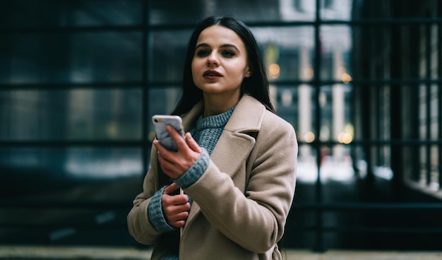 Giovane bruna alla moda con il trucco e in caldo cappotto beige che tiene il telefono cellulare