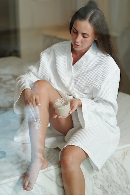 Giovane bruna che applica una crema per il corpo nutriente sulle gambe mentre è seduta su un tavolo di marmo in hamam