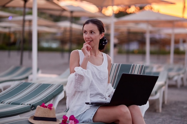 Giovane bruna con un laptop su un lettino sulla spiaggia Freelancer che lavora a distanza webinar concetto di apprendimento in chat con gli amici Nuovo concetto normale