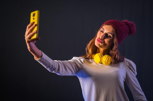 Giovane brunetta caucasica con cuffie gialle, maglietta bianca e cappello di lana su sfondo nero, facendo un selfie con il cellulare