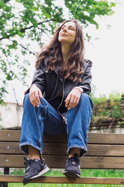 Giovane brunetta in giacca di pelle e jeans Una giovane modella positiva e allegra è seduta su una panchina e posa per la fotocamera