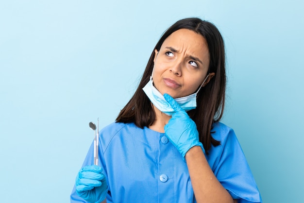 Giovane brunetta razza mista dentista donna che tiene gli strumenti sopra la parete isolata avendo dubbi mentre cerca