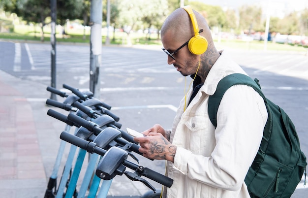 Giovane calvo con cuffie gialle che utilizza un'app per telefono cellulare per guidare uno scooter pubblico in città