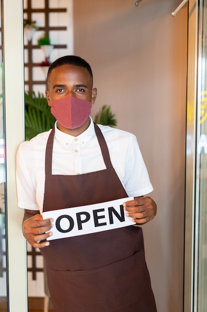 Giovane cameriere africano in uniforme e maschera protettiva in possesso di carta con avviso aperto mentre si trova davanti alla porta aperta del bar o del ristorante