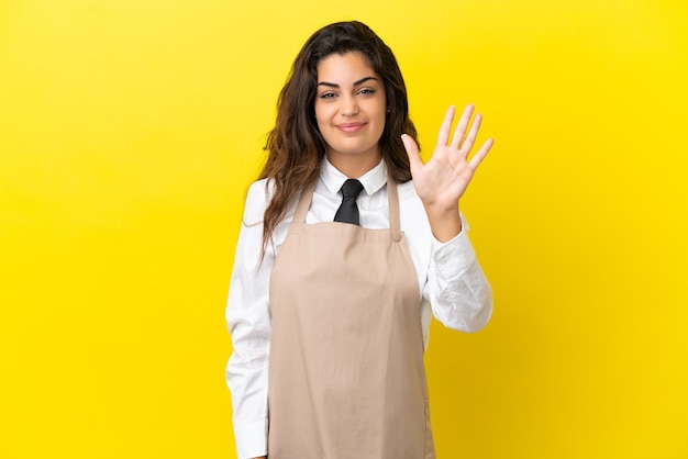 Giovane cameriere caucasico del ristorante isolato su sfondo giallo contando cinque con le dita