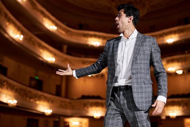 Giovane cantante attraente di talento in elegante abito classico, cantando hit con la bocca aperta nel gesto del microfono con la mano in piedi sul palco del teatro. Bel ragazzo caucasico durante l'esibizione