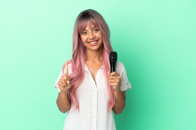 Giovane cantante di razza mista con i capelli rosa isolati su sfondo verde che mostra e solleva un dito