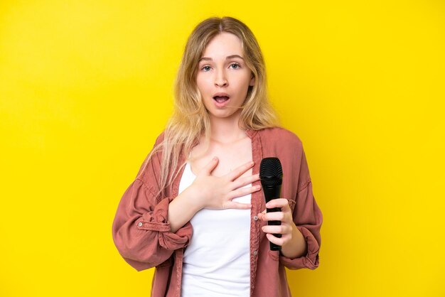 Giovane cantante donna caucasica che prende in mano un microfono isolato su sfondo giallo sorpreso e scioccato mentre guarda a destra