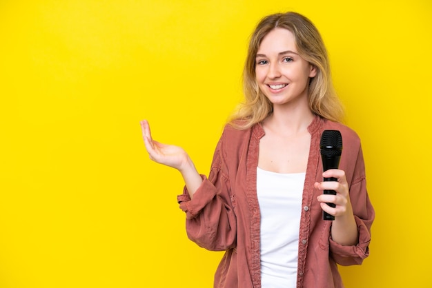 Giovane cantante donna caucasica che prende un microfono isolato su sfondo giallo che estende le mani di lato per invitare a venire