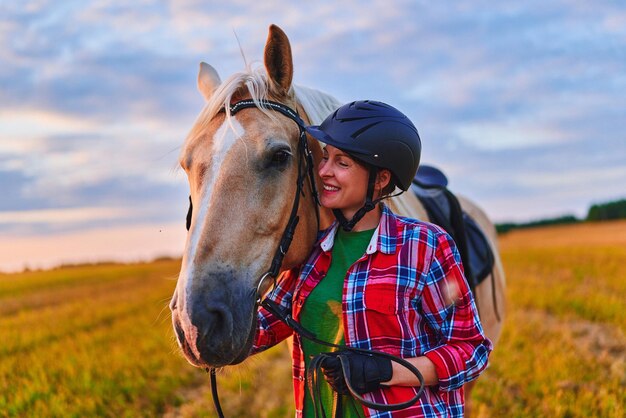 Giovane carino felice gioioso soddisfatto sorridente donna pilota che indossa il casco abbracciando e accarezzando bellissimo cavallo biondo palomino al prato al tramonto all'ora d'oro