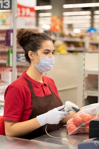 Giovane cassiera bruna in uniforme, guanti protettivi e confezione di scansione maschera di pomodori freschi maturi al bancone del supermercato