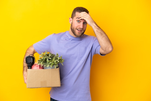 Giovane caucasico che fa una mossa mentre prende in mano una scatola piena di cose isolate sul muro giallo che fa il gesto di sorpresa mentre guarda al lato