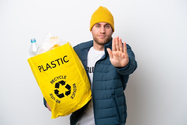 Giovane caucasico che tiene un sacchetto pieno di bottiglie di plastica da riciclare isolato su sfondo bianco facendo un gesto di arresto