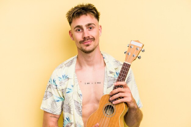 Giovane caucasico che tiene un ukulele isolato su sfondo giallo