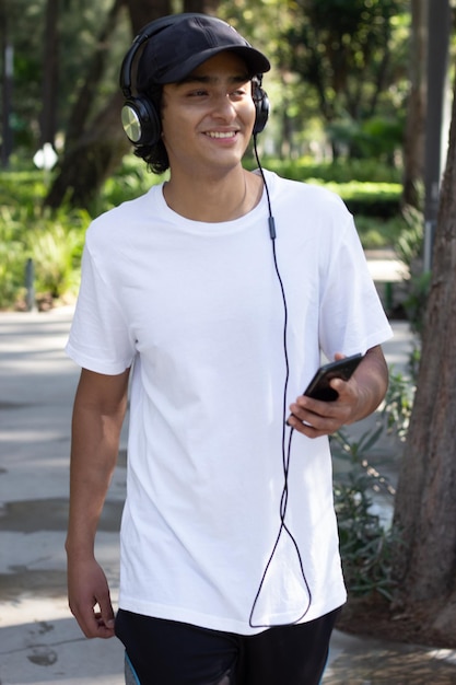 Giovane che ascolta musica con le cuffie mentre passeggia nel parco sorridente e rilassato in mezzo alla natura, indossando un berretto e una maglietta bianca