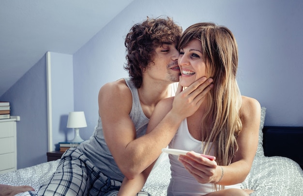 Giovane che bacia una bella donna mentre mostra il test di gravidanza in camera da letto