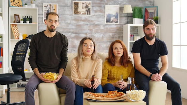 Giovane che batte le mani mentre guarda un film con i suoi amici in soggiorno mangiando pizza e bevendo birra.