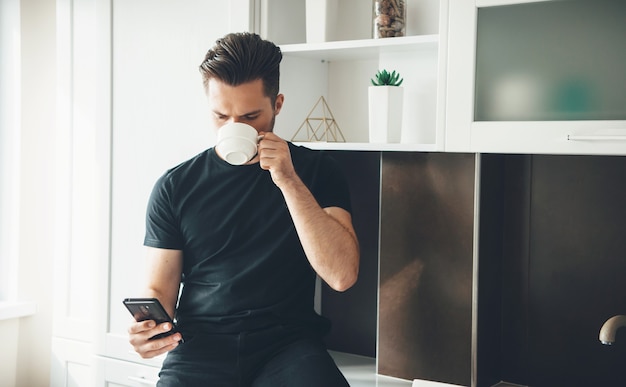 Giovane che beve un caffè in cucina mentre chiacchiera sul cellulare che indossa abiti neri