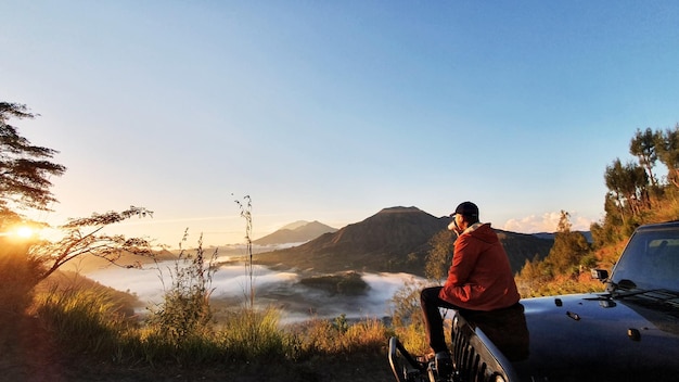 Giovane che beve una tazza di bevanda calda e si siede sul cofano dell'auto mentre si gode la vista sulle montagne di Batur all'ora dell'alba