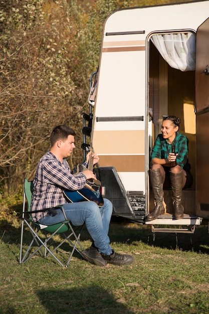 Giovane che cerca di cantare una canzone alla chitarra per la sua ragazza davanti al loro camper retrò. Coppia allegra.