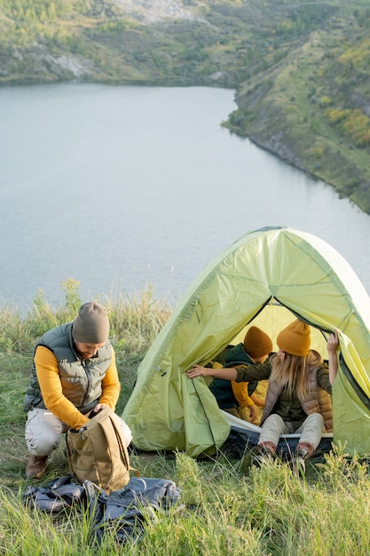 Giovane che cerca qualcosa nello zaino mentre si accovaccia sull'erba verde vicino alla tenda dove sua moglie e suo figlio riposano durante il viaggio nel fine settimana