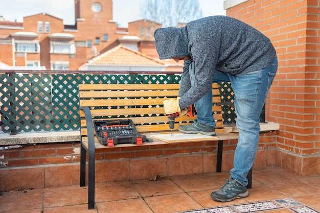 Giovane che fa il bricolage sulla terrazza di casa sua con un trapano a batteria su una panca di legno