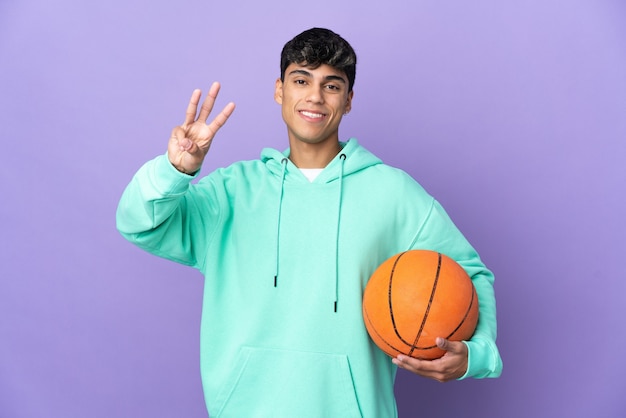Giovane che gioca a basket su sfondo viola isolato felice e contando tre con le dita