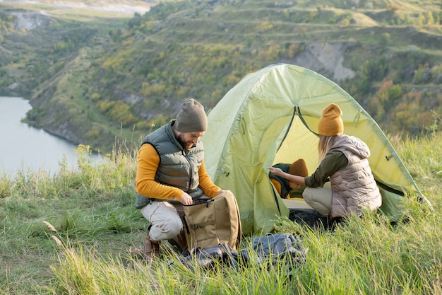 Giovane che guarda nello zaino mentre si accovaccia sull'erba verde vicino alla tenda dove sua moglie e suo figlio riposano durante il viaggio nel fine settimana autunnale