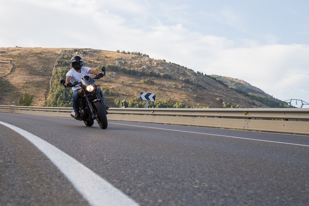 Giovane che guida un motociclo sulla strada che gira una curva nelle montagne il giorno soleggiato.