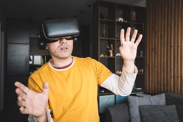 Giovane che indossa cuffie o occhiali per realtà virtuale in piedi a casa guardandosi intorno e cercando di toccare qualcosa di eccitato dall'effetto partecipazione guardando film 3d vr per telefono e giochi mobili