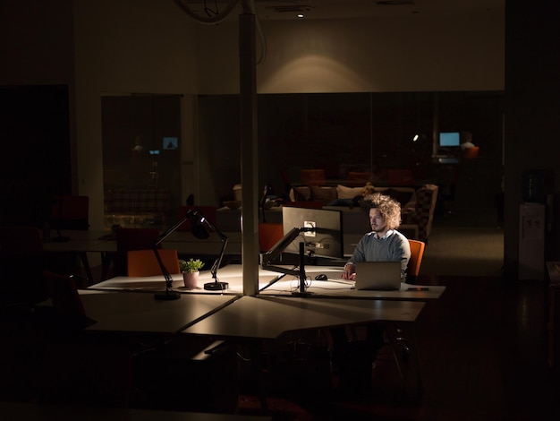 Giovane che lavora al computer di notte in un ufficio buio. Il designer lavora in un secondo momento.