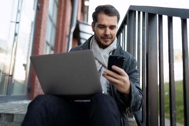 Giovane che lavora al computer portatile e guarda lo schermo del telefono cellulare mentre è seduto sui gradini all'esterno