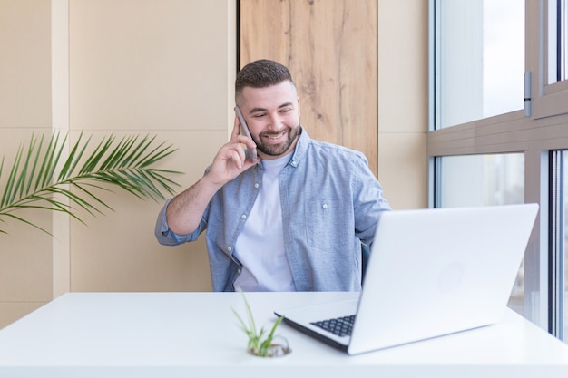 Giovane che lavora dall'ufficio seduto vicino alla finestra al laptop vestito con abiti casual