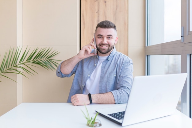 Giovane che lavora dall'ufficio seduto vicino alla finestra al laptop vestito con abiti casual