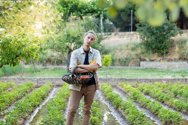 Giovane che lavora in giardino con le braccia incrociate mentre tiene un cesto di verdure e il campo sullo sfondo