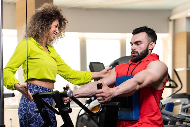 Giovane che lavora in palestra facendo esercizio per la schiena sulla macchina con l'aiuto del suo allenatore